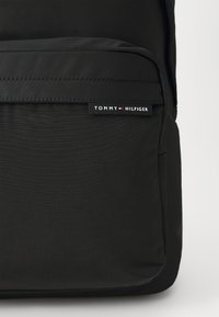 Fekete hátizsák sima külsővel, elöl egy zsebbel és egy felirattal, melyen a "TOMMY HILFIGER" fehér és piros szöveggel olvasható.