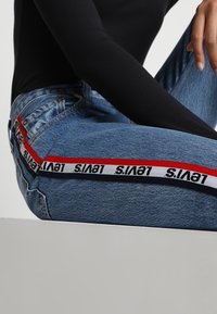 Jeans in denim azzurro chiaro con una fascia a righe rossa e bianca con il marchio "Levi's" lungo il lato, abbinati a una maglietta nera a maniche lunghe.
