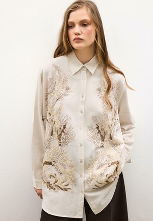 Giovane donna che indossa una camicia beige con bottoni e un ricamo floreale intricato beige e marrone, in piedi davanti a uno sfondo chiaro e semplice.