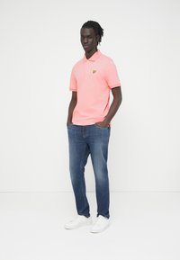 Φωτεινό ροζ πόλο πουκάμισο με κοντά μανίκια και κίτρινο λογότυπο, συνδυασμένο με μπλε τζιν και λευκά αθλητικά παπούτσια. Λείο ύφασμα, καθημερινό σχέδιο.