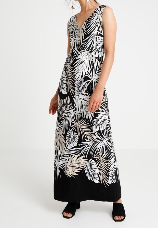 Femme portant une robe maxi noire sans manches avec un imprimé feuilles tropicales blanches, associée à des sandales noires à talons ouvertes.