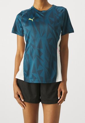 Persona vistiendo una camiseta de Puma de patrón geométrico en teal oscuro con paneles laterales blancos y pantalones cortos negros, de pie frente a un fondo liso.