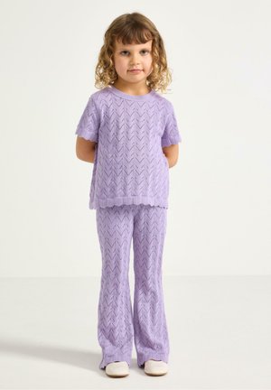 Nuori tyttö, jolla on kiharat hiukset ja joka pukeutunut yhteensopivaan laventelinväriseen neulepaitaan ja -housuihin, seisoo käsivartensa selän takana yksiväristä taustaa vasten.