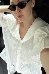 Bela bomba iz bombaža s detajli iz stopenj, napihnjenimi rokavi in okroglo ovratnico. Ima zapiranje na gumb in zapletene čipkaste poudarke.