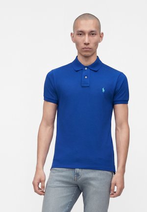 Homme au crâne rasé portant un polo bleu à manches courtes et un jean bleu clair, debout devant un fond blanc uni.