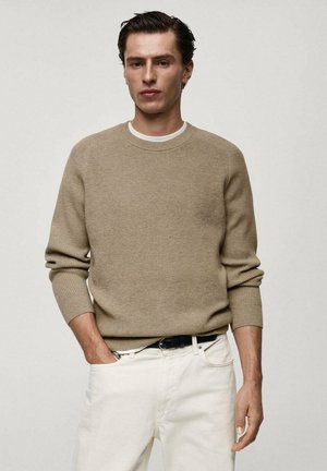 Beige knitted sweater with ribbed texture, round neckline, and long sleeves, paired with white jeans and a black belt.