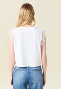 Valkoinen puuvillapaita, jossa on pitsireunus olkapäillä, lyhyt malli ja yksinkertainen pyöreä kaula-aukko. Yhdistetty sinisiin korkeavyötäröisiin farkkuihin.