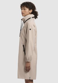 Cappotto impermeabile beige con colletto alto, maniche elastiche, tasche laterali con zip e dettagli in metallo nero. Texture liscia, design fino al ginocchio.