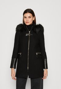 Cappotto nero con cappuccio in pelliccia sintetica, chiusura a zip e dettagli in metallo argentato. Presenta maniche effetto pelle e due tasche zip frontali.