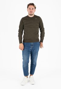 Maglione lungo a maniche imbottite di colore verde scuro con texture a coste abbinato a jeans tapered blu chiaro e sneakers bianche, in piedi su uno sfondo bianco.