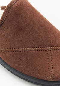 Bruine suède pantoffel met een gladde textuur, voorzien van contrasterende stiksels en een zwarte rubberen zool. Rondvormig met zachte randen.