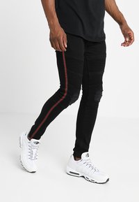 Jeans skinny negros con una franja lateral roja, que presentan detalles desgastados y secciones acanaladas. Combinados con zapatillas blancas.