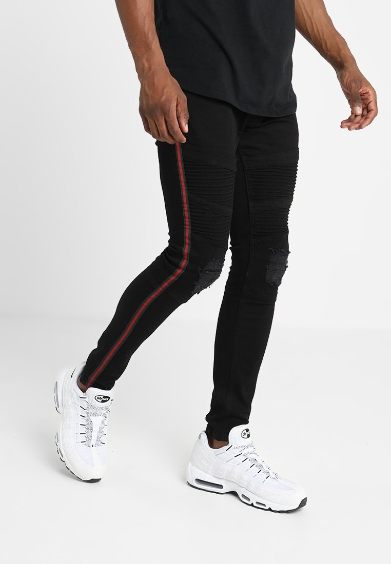 Jeans skinny negros con una franja lateral roja, que presentan detalles desgastados y secciones acanaladas. Combinados con zapatillas blancas.