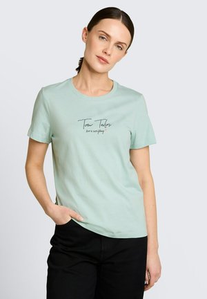 Kobieta o brązowych włosach związanych w kucyk, ubrana w jasnuzołtą koszulkę Tom Tailor z napisem "miłość to wszystko", zestawioną z czarnymi spodniami.