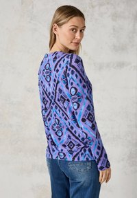 Paarse en blauwe gemusterde trui met een geometrisch ontwerp, voorzien van een ronde hals en lange mouwen, gecombineerd met blauwe spijkerbroeken.