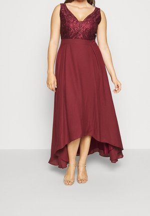 Bordó šaty bez rukávů s krajkovým živůtkem, výstřihem do V, přiléhavým pasem a sukní s vysokým předním a nízkým zadním okrajem, nošené s béžovými podvazkovými botami na podpatku.