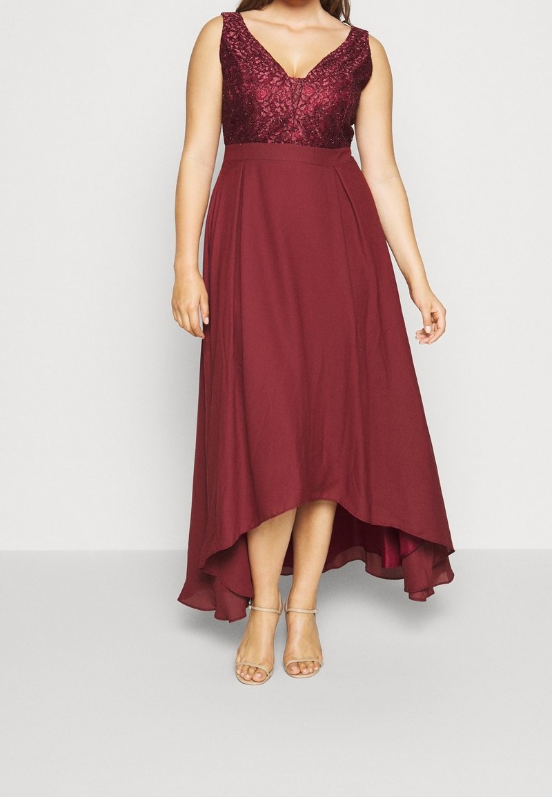 Robe sans manches bordeaux avec corsage en dentelle, encolure en V, taille cintrée et jupe asymétrique, portée avec des talons beiges à bride autour de la cheville.
