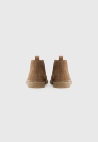 Botines de ante marrón con punta redonda, paneles laterales elásticos y un tacón pequeño. Presentan una lengüeta en la parte trasera para facilitar su uso.