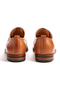 Scarpe in pelle marrone con una texture liscia, tacco arrotondato e una striscia nera a contrasto sulla suola in gomma. Presentano un design slip-on.