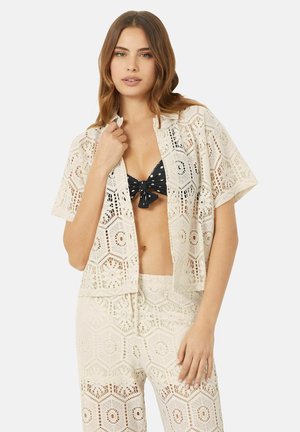 Couverture en dentelle crochetée avec des motifs géométriques, à manches courtes et à ouverture sur le devant, de couleur crème clair, portée sur un haut de bikini à pois noirs.