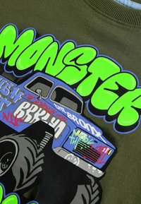 Grüner "MONSTER" Text über einem blauen Monstertruck mit großen Reifen und Graffiti-Schrift auf olivgrünem Stoffhintergrund.