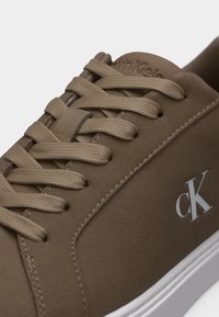 Καφέ αθλητικό παπούτσι Calvin Klein με ομοιόχρωμους κορδόνια, λευκό λογότυπο "cK" στο πλάι και λευκή ελαστική σόλα, εμφανιζόμενο σε κοντινή λήψη.