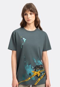 Grijze katoenen T-shirt met een spatschilderijgrafiek, een schilderkwast en gele "NAT VERF" waarschuwingslintprint aan de onderkant.