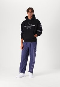 Sort hættejakke med teksten "LOVE SCARS", lilla detaljer og et stjernedesign. Matchet med marineblå joggers med hvide kanter og logoer. Hvide sneakers.