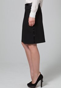 Jupe noire avec un ourlet évasé, présentant un panneau latéral en satin. Associée à des escarpins noirs en daim et un chemisier blanc.