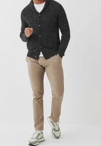 Gruby czarny sweter z dzianiny z kołnierzem typu szal, brązowymi guzikami, noszony na białej koszuli i zestawiony z beżowymi spodniami o slim fit oraz szarymi trampkami.