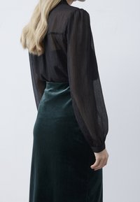 Svart genomskinlig blus med puffärmar, i kombination med en mörkgrön sammetskirt. Tyget har en subtil glans och en mjuk textur.