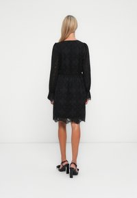 Eine Frau mit langen blonden Haaren steht mit dem Rücken zu uns und trägt ein knielanges schwarzes Spitzenkleid sowie schwarze High Heels.