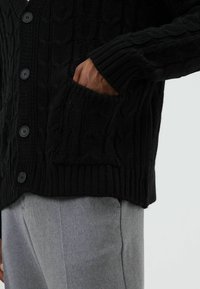 Zwarte gebreide cardigan met een getextureerd kabelpatroon, voorzien van grote zwarte knopen en een zijvak. Draagt over grijze broek.