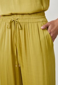 Pantalones de un suave verde oliva claro con cinturilla elástica y lazo ajustable. Tejido suave y liso, con bolsillos laterales y un sutil drapeado.