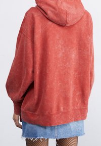 Sudadera roja de gran tamaño con acabado texturizado, capucha y puños acanalados, combinada con una minifalda de mezclilla clara y medias de red.