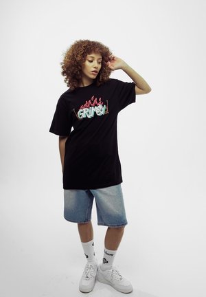 Camiseta de algodón negra con el logo "GRIMY" en llamas color azul y rosa, combinada con pantalones cortos de mezclilla y zapatillas blancas, sobre un fondo simple.