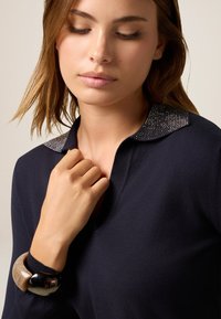 Marine pullover met een V-hals en een gestructureerde kraag met decoratieve stenen. Een grote, contrasterende armband siert de pols.