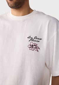 White cotton t-shirt with a crew neck, featuring black script text "Les Deux Fleurs" and a pink floral embroidery on the left chest.