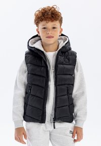 Svart huva puffer väst med quiltad struktur, frontdragkedja och sidofickor, bärs över en ljusgrå sweatshirt och matchande sweatpants.