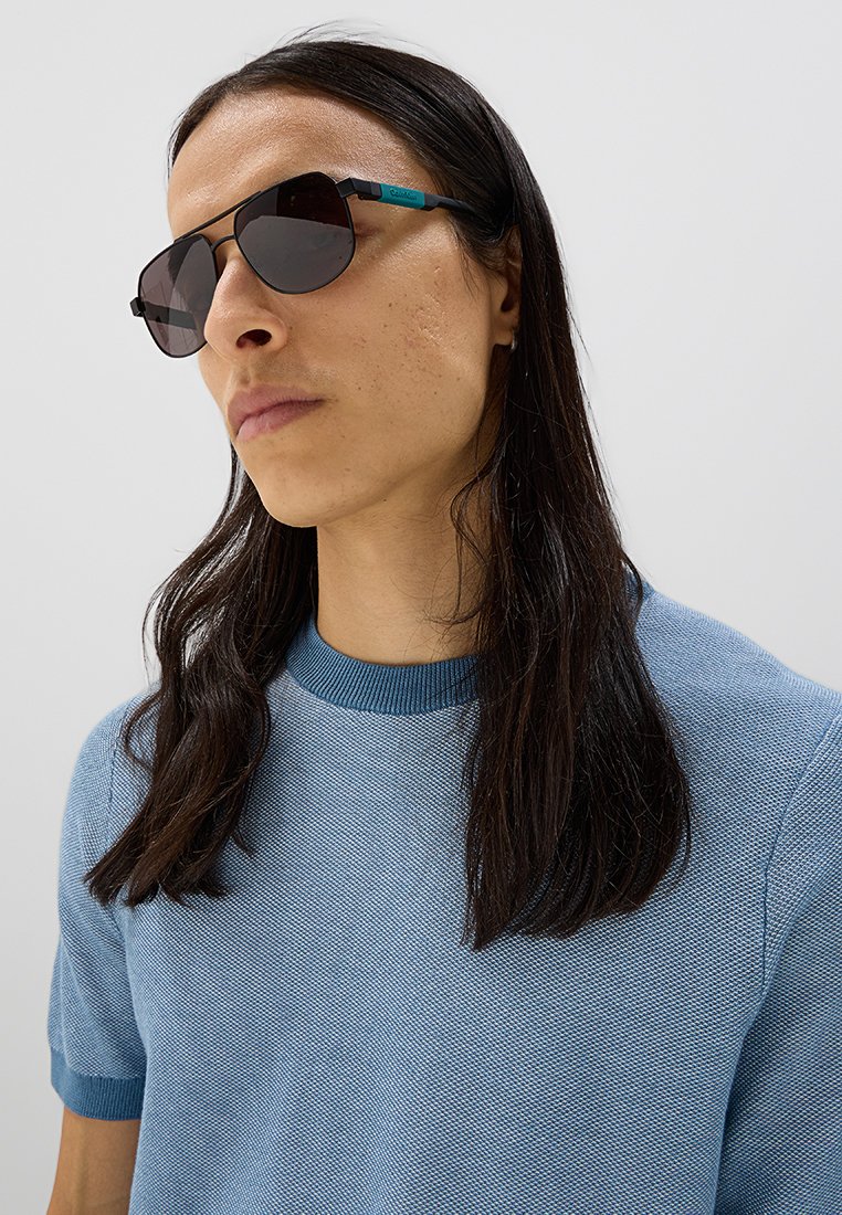 Gafas de sol con montura negra y lentes oscuros, con un acento azul en las patillas. Llevadas con una camisa azul texturizada.