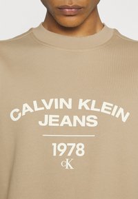 Felpa beige con la scritta "CALVIN KLEIN JEANS" e "1978" in bold bianco, collo rotondo e materiale in cotone morbido.