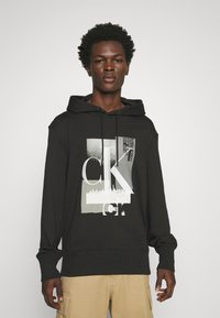 Felpa nera con cappuccio caratterizzata da un design grafico in bianco e grigio, con un aspetto a strati e le lettere "CK" in evidenza.