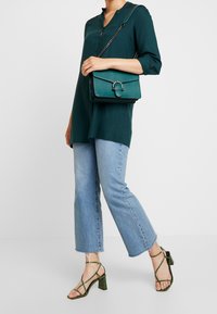 Kvinde iført en teal-bluse, lyseblå cropped jeans, grønne sandaletter med blokhæl og bærende en matchende grøn lædertaske.