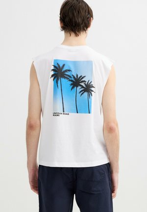 Persona que lleva una camiseta blanca sin mangas con un gráfico cuadrado azul de tres palmeras y el texto "Lincoln Road Miami" en la espalda.