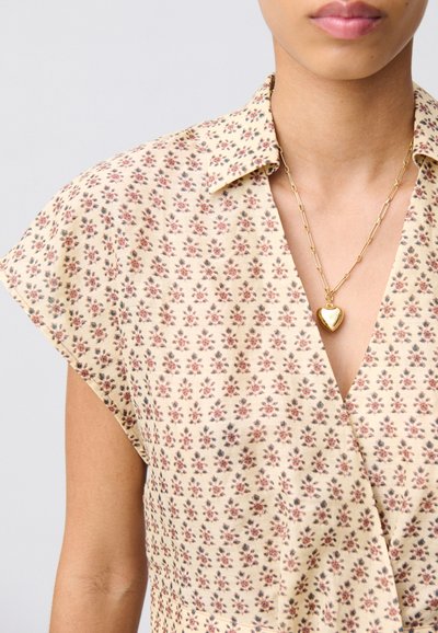 Blusa floreale in crema con maniche a cappuccio, caratterizzata da un piccolo motivo di fiori rossi e blu. Una collana con pendente a cuore dorato completa il design.