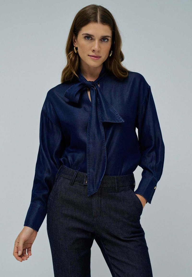 Blusa azul marino de tela de mezclilla con diseño de cuello atado, manga larga y un ligero brillo. Combinada con pantalones de mezclilla oscuros que tienen una textura suave.