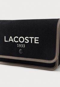 Fekete szövet tasak bézs díszítésekkel, fehér "LACOSTE 1933" logóval. Nyomógombos zárással és varrott szélekkel készült.