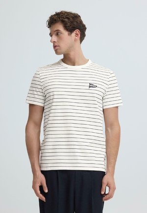 Junger Mann mit lockigem Haar, der ein weißes T-Shirt mit dünnen schwarzen horizontalen Streifen und dunkle Hosen trägt, schaut nach links.