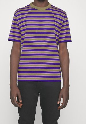 Mand iført en kortærmet t-shirt med striber i lilla og olivengrøn kombineret med sorte bukser, stående mod en ensfarvet baggrund.