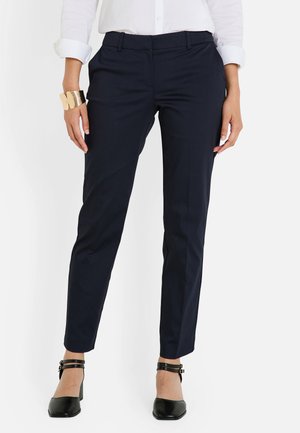 Femme portant un pantalon ajusté bleu marine, une chemise blanche à manches retroussées, des escarpins noirs et un large bracelet en or au poignet gauche.